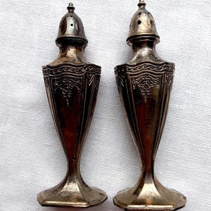 May include: A pair of antique silver salt and pepper shakers. The shakers have a decorative, ornate design with a flared base and a domed top with small holes. The metal has a tarnished appearance, suggesting age.
