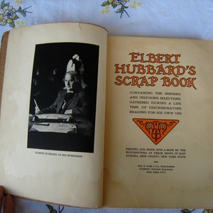 May include: An open book titled Elbert Hubbard's Scrap Book with a black and white photograph of Elbert Hubbard in his workshop on the left page and printed text on the right page, including details about the Roycrofters and the publisher.