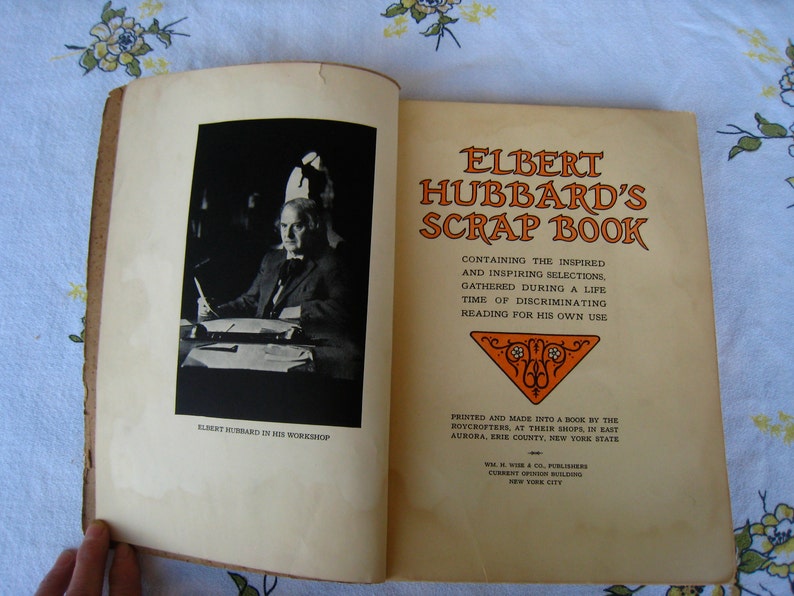 May include: An open book titled Elbert Hubbard's Scrap Book with a black and white photograph of Elbert Hubbard in his workshop on the left page and printed text on the right page, including details about the Roycrofters and the publisher.