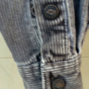 May include: Close-up of a gray corduroy shirt with white stripes. The shirt has a button-up front and a long sleeve. The buttons are metal and have a raised design.