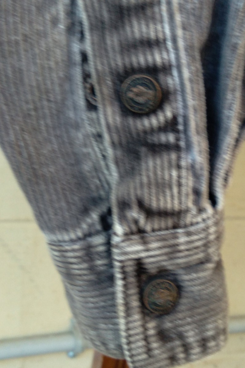May include: Close-up of a gray corduroy shirt with white stripes. The shirt has a button-up front and a long sleeve. The buttons are metal and have a raised design.