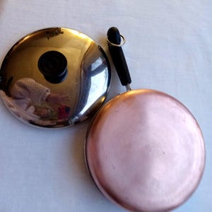 May include: A copper-bottomed frying pan with a black handle and a stainless steel lid. The lid reflects the surroundings, including a person's arm and a red object. The pan is round and has a shiny copper surface.