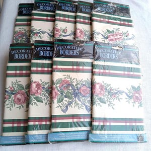 May include: A collection of decorative borders in plastic packaging. Each roll features a floral design with pink roses, blue ribbons, and green foliage against a cream background. The borders have green and burgundy stripes.