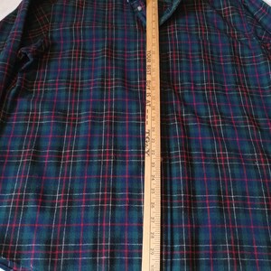May include: A long-sleeve button-down shirt featuring a dark green, navy blue, and red plaid pattern. A wooden yardstick is placed vertically over the center, showing measurements up to 32 inches with the text YOUR BEST BUY IS AT TG&Y.