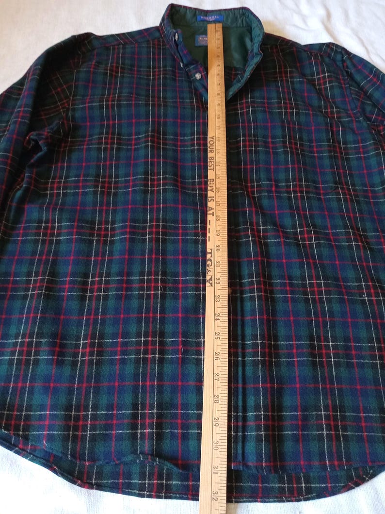May include: A long-sleeve button-down shirt featuring a dark green, navy blue, and red plaid pattern. A wooden yardstick is placed vertically over the center, showing measurements up to 32 inches with the text YOUR BEST BUY IS AT TG&Y.