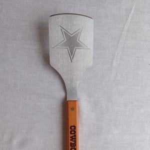 May include: A stainless steel spatula with a wooden handle. The spatula features a large star design etched into the metal. The handle is engraved with the word "COWBOYS" in bold, vertical letters.