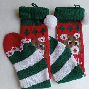 May include: Christmas stockings and matching mittens. The stockings are red with a reindeer and tree pattern, green cuffs, and white pom-poms. The mittens are red with a white and green striped pattern.