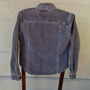 May include: A gray corduroy shirt with a button-down collar and long sleeves. The shirt has a faded look and is hanging on a wooden hanger.