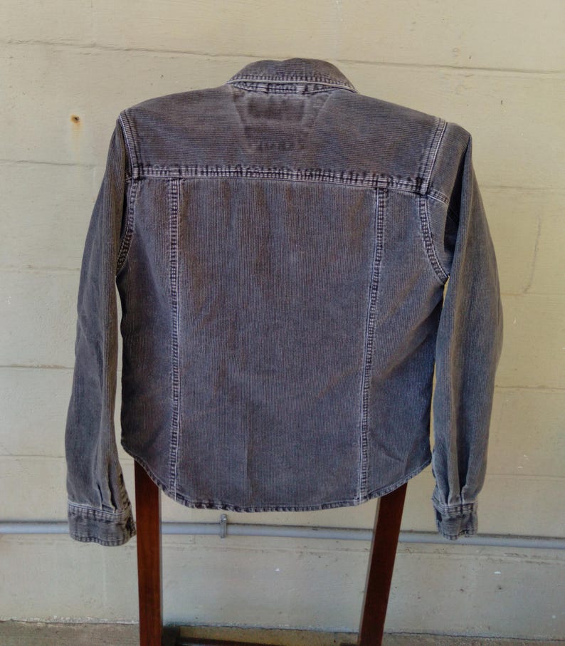 May include: A gray corduroy shirt with a button-down collar and long sleeves. The shirt has a faded look and is hanging on a wooden hanger.