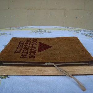 May include: A rectangular scrapbook with a textured tan cover featuring the brown printed text Elbert Hubbard's Scrap Book and a red triangular emblem. The book is bound with a thin, light-colored cord tied at the front edge, and the pages are visible.