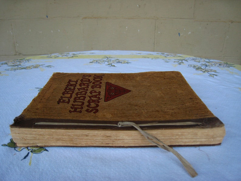 May include: A rectangular scrapbook with a textured tan cover featuring the brown printed text Elbert Hubbard's Scrap Book and a red triangular emblem. The book is bound with a thin, light-colored cord tied at the front edge, and the pages are visible.