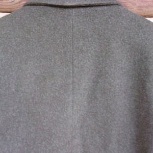 May include: Close-up of the back of a gray wool blazer. The blazer has a notched collar and a center back seam. The fabric has a slightly textured appearance. The background is a wooden surface.