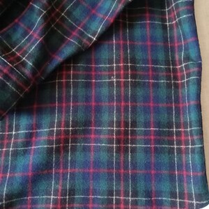 May include: A fabric featuring a classic tartan plaid pattern with a deep green base, intersecting navy blue stripes, and thin, vibrant red and white lines forming a grid. The textile has a soft, slightly fuzzy texture throughout the surface.