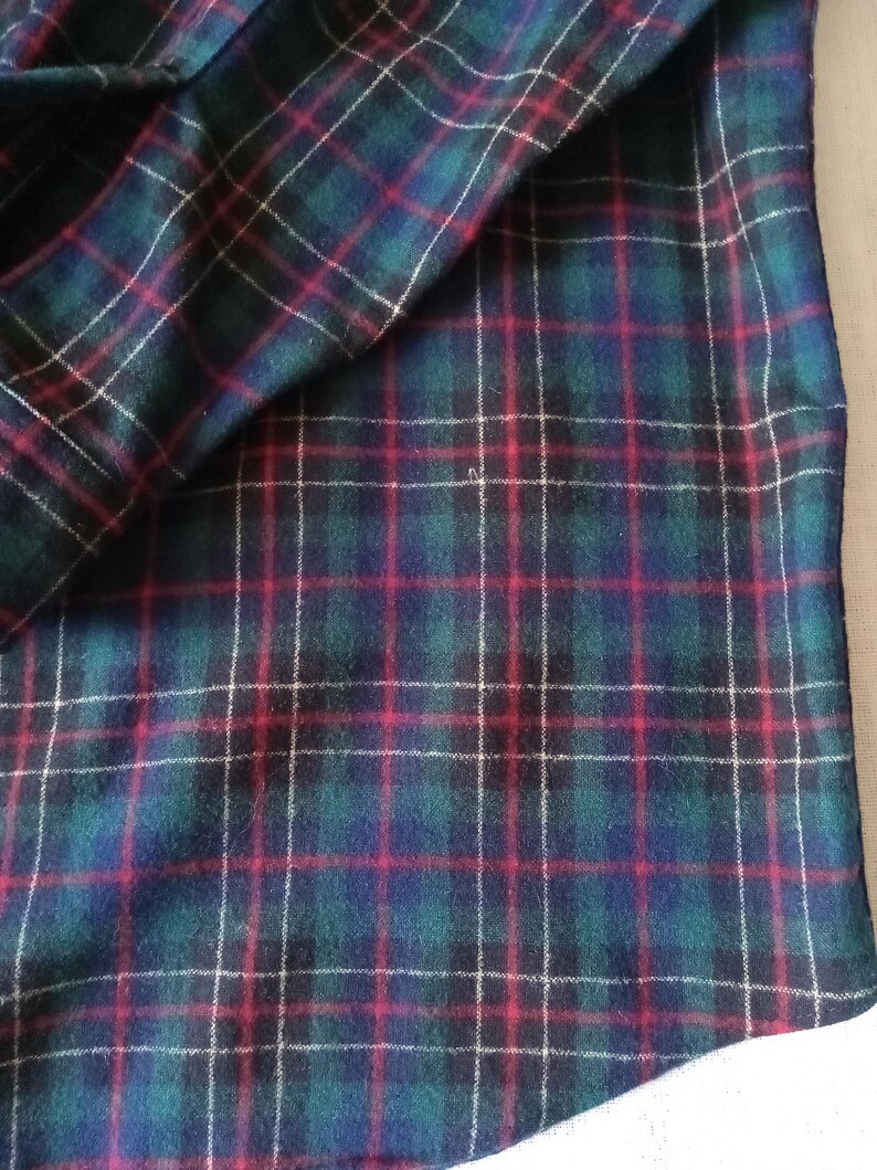 May include: A fabric featuring a classic tartan plaid pattern with a deep green base, intersecting navy blue stripes, and thin, vibrant red and white lines forming a grid. The textile has a soft, slightly fuzzy texture throughout the surface.