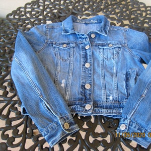 May include: A light blue denim jacket with a classic button-up design. The jacket features two chest pockets, silver buttons, and distressed detailing. The sleeves are long with buttoned cuffs. The jacket is a cropped style.