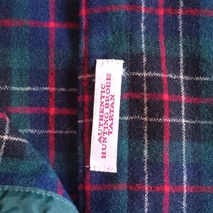May include: A fabric with a dark blue, green, and red plaid tartan pattern. A white rectangular tag is sewn onto the fabric, featuring red text that reads AUTHENTIC HUNTING BRODE TARTAN. The material has a soft, textured appearance.
