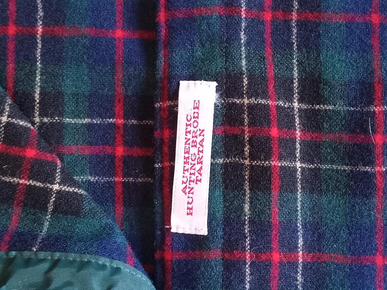 May include: A fabric with a dark blue, green, and red plaid tartan pattern. A white rectangular tag is sewn onto the fabric, featuring red text that reads AUTHENTIC HUNTING BRODE TARTAN. The material has a soft, textured appearance.
