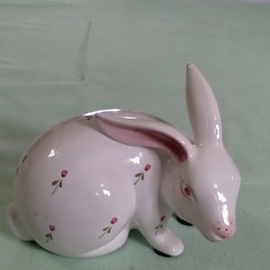 May include: A white ceramic rabbit figurine with pink ears and painted pink flowers. The rabbit is sitting on a green surface.