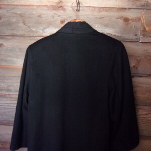 May include: A black blazer with a long, open collar. The blazer is hanging on a wooden hanger against a wood panel background.