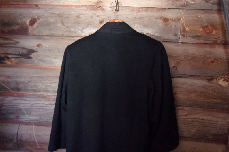 May include: A black blazer with a long, open collar. The blazer is hanging on a wooden hanger against a wood panel background.