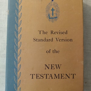 May include: The cover of a blue and brown book titled "The Revised Standard Version of the NEW TESTAMENT". A cross with a book underneath is in the center of the cover.