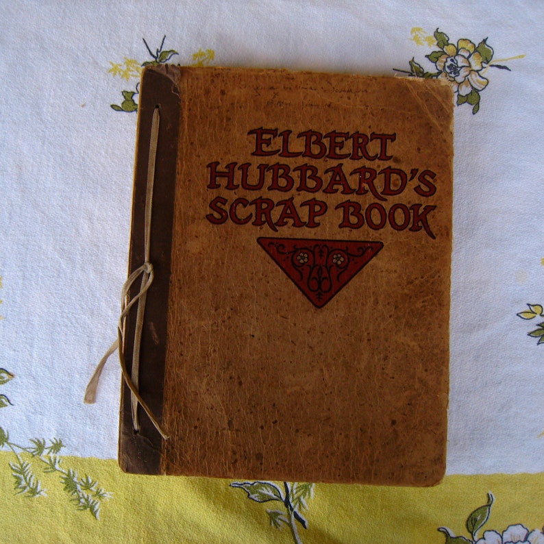 May include: A vintage tan scrap book titled ELBERT HUBBARD'S SCRAP BOOK in dark red lettering. The cover features a dark red triangular decorative element. The book is bound with a dark brown spine and a light-colored cord tied in a knot.
