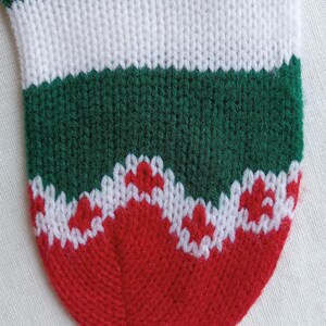 May include: Close-up of a knitted Christmas stocking. The stocking features alternating bands of green and white, with a red toe and a white and red patterned design near the toe. The knit texture is clearly visible.