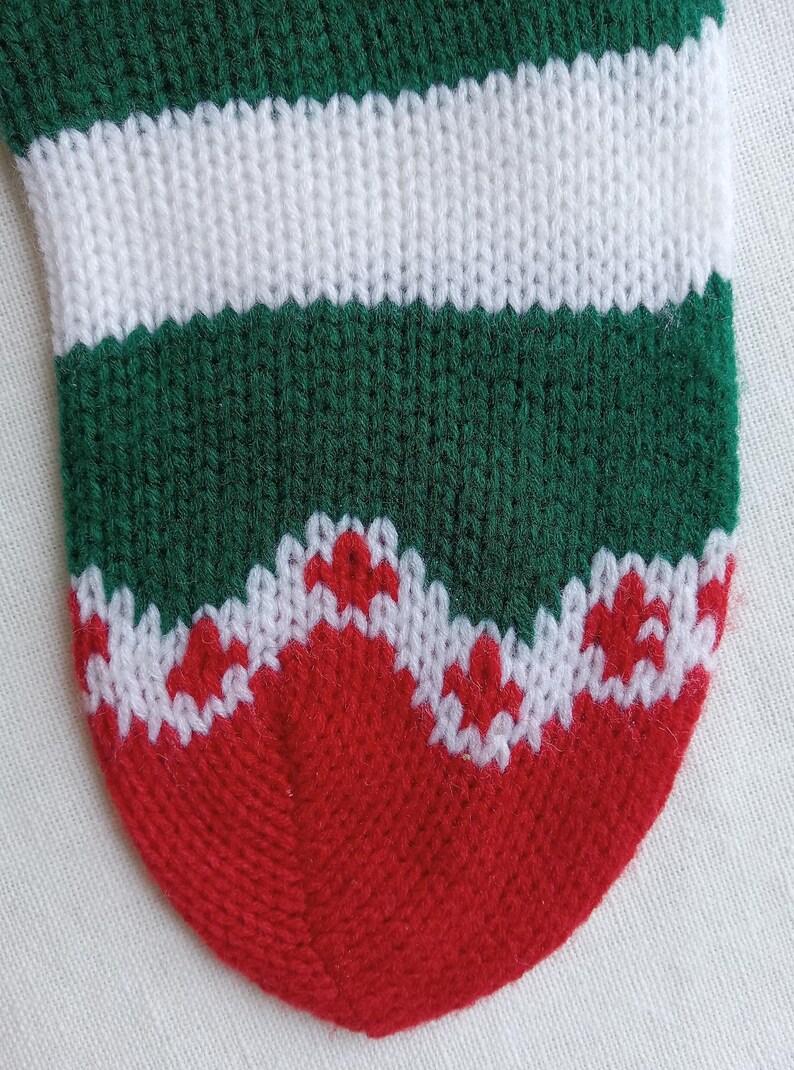 May include: Close-up of a knitted Christmas stocking. The stocking features alternating bands of green and white, with a red toe and a white and red patterned design near the toe. The knit texture is clearly visible.