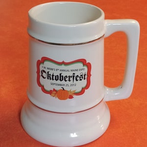 May include: White ceramic beer stein with a gold rim and a red, green, and white logo that says "F.W. Webb's 9th Annual Maine Expo Oktoberfest September 25, 2012".