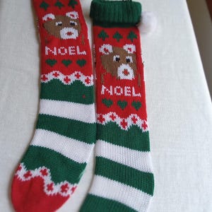 May include: Two Christmas stockings with a red, green, and white color scheme. Each stocking features a knitted design with a bear, the word "NOEL", and decorative patterns. A white pom-pom is attached to the top.