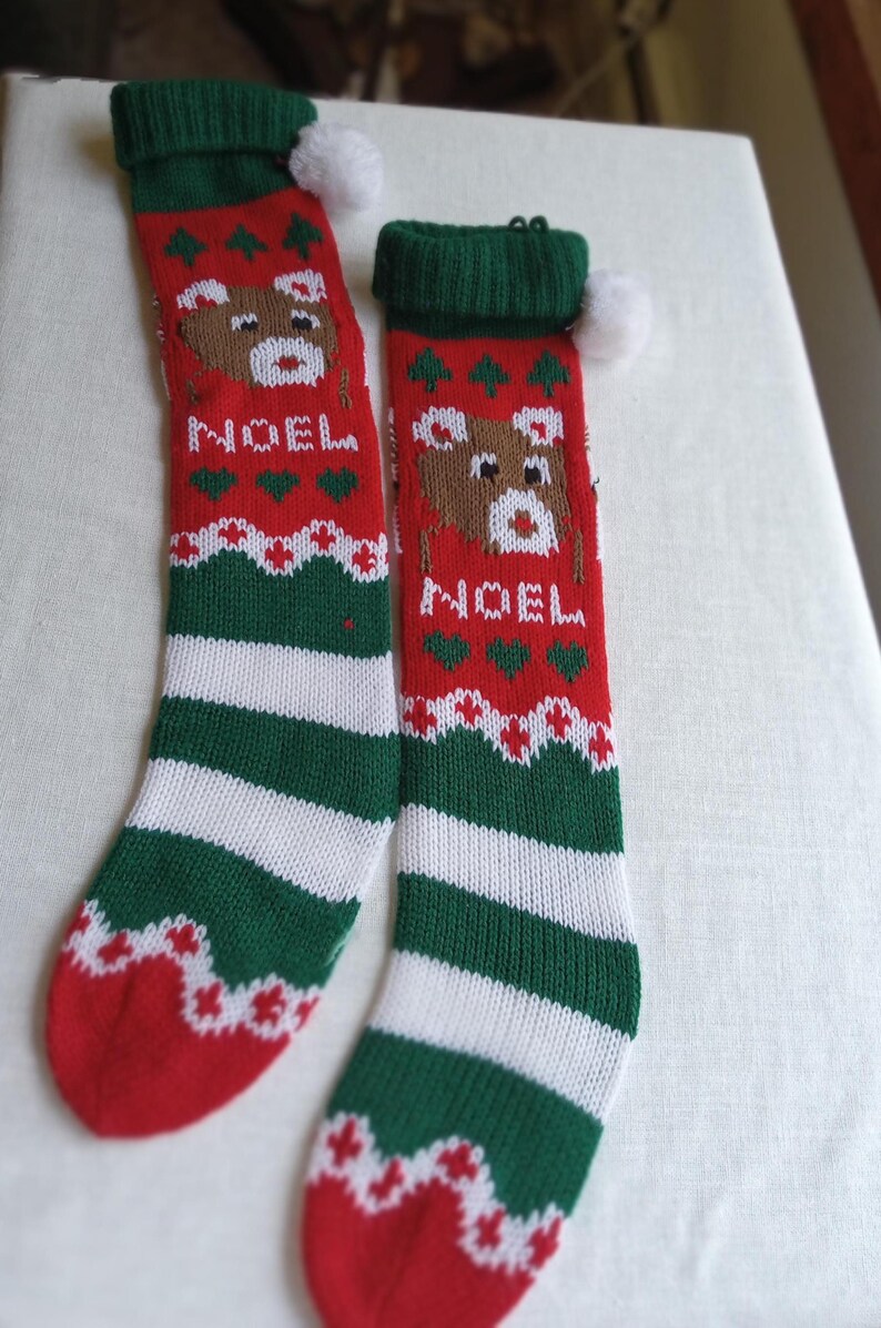 May include: Two Christmas stockings with a red, green, and white color scheme. Each stocking features a knitted design with a bear, the word "NOEL", and decorative patterns. A white pom-pom is attached to the top.