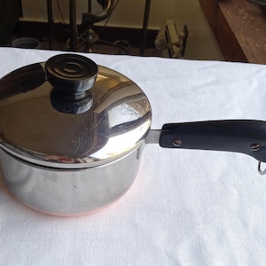 May include: A stainless steel saucepan with a lid and a black handle. The pot has a copper-colored bottom and a black knob on the lid. The handle has a metal loop at the end. The pot is sitting on a white surface.