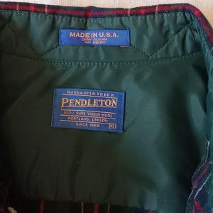 May include: A dark green quilted interior lining of a garment with a red, green, and blue plaid collar. Two blue labels are stitched inside: one reads MADE IN U.S.A. DRY CLEAN, and the other reads WARRANTED TO BE A PENDLETON 100% PURE VIRGIN WOOL.