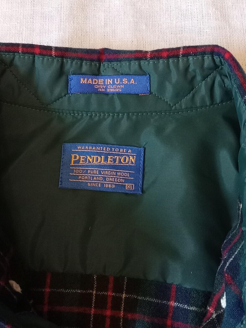 May include: A dark green quilted interior lining of a garment with a red, green, and blue plaid collar. Two blue labels are stitched inside: one reads MADE IN U.S.A. DRY CLEAN, and the other reads WARRANTED TO BE A PENDLETON 100% PURE VIRGIN WOOL.
