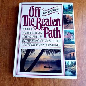 May include: A hardcover book titled "Off The Beaten Path" by Reader's Digest. The cover features a scenic lake image with a canoe and text in burgundy and white. Additional images of landscapes are on the right side.