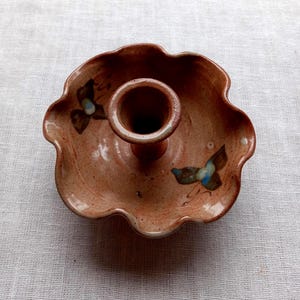 May include: A terracotta-colored ceramic candle holder with a scalloped, flower-like rim. The surface features a speckled glaze and two hand-painted butterfly motifs in shades of brown and blue. The central cylindrical socket is raised.