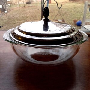 May include: A clear glass serving bowl with a silver lid. The lid has a black finial and engraved initials. The bowl sits on a wooden surface, with a window and outdoor scene in the background.