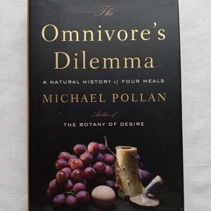 May include: A black book cover with gold lettering that reads "The Omnivore's Dilemma: A Natural History of Four Meals" by Michael Pollan, author of "The Botany of Desire". The cover features a still life of grapes, an egg, and a bone.