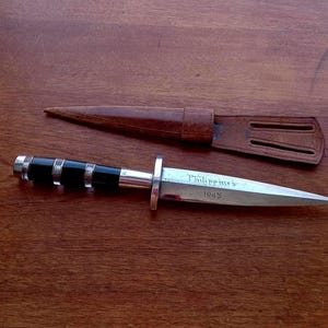 May include: A vintage dagger with a silver blade and a black and silver handle, accompanied by a brown leather sheath. The blade is inscribed with "Philippines 1945". The dagger is displayed on a wooden surface.