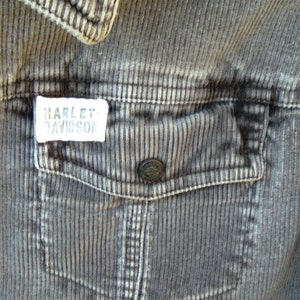 May include: A close-up of a gray corduroy jacket with a patch that reads "Harley Davidson" on the left side of the chest. The jacket has a large pocket on the right side of the chest.