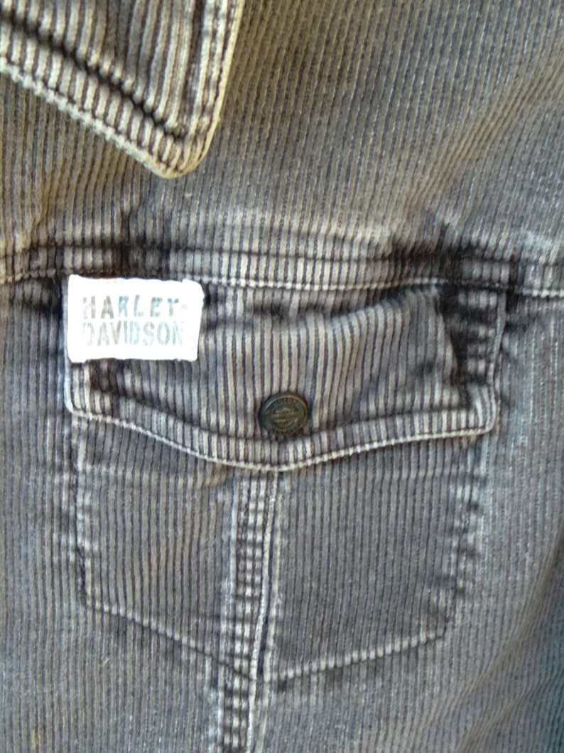 May include: A close-up of a gray corduroy jacket with a patch that reads "Harley Davidson" on the left side of the chest. The jacket has a large pocket on the right side of the chest.