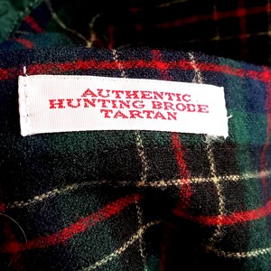 May include: A fabric featuring a dark green, black, and red plaid pattern with a white rectangular label stitched onto the surface. The label contains red text that reads AUTHENTIC HUNTING BRODE TARTAN in a clear, capitalized font style.