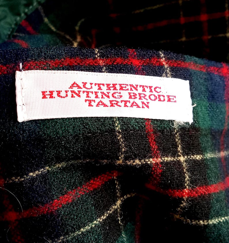 May include: A fabric featuring a dark green, black, and red plaid pattern with a white rectangular label stitched onto the surface. The label contains red text that reads AUTHENTIC HUNTING BRODE TARTAN in a clear, capitalized font style.