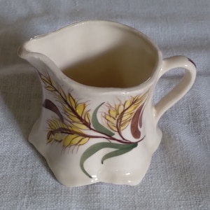 May include: A white ceramic creamer with a ruffled edge and a handle. The creamer is decorated with a yellow and brown wheat design.