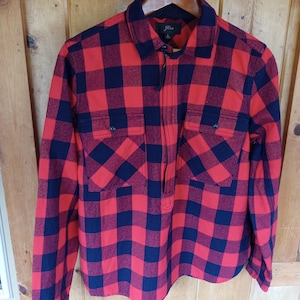 May include: Red and navy blue plaid shirt with two chest pockets and a half-zip closure. The long-sleeved shirt is hanging on a wooden hanger against a wooden background. The shirt is from J.Crew.