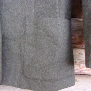 May include: Close-up of a dark olive green blazer. The blazer has a single button closure with a decorative button. The blazer has a pocket on the front and a sleeve is visible. The fabric appears to be a wool blend.