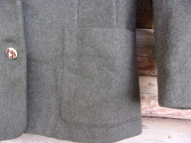 May include: Close-up of a dark olive green blazer. The blazer has a single button closure with a decorative button. The blazer has a pocket on the front and a sleeve is visible. The fabric appears to be a wool blend.