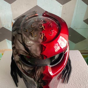 Puede incluir: Un casco de motocicleta personalizado con un diseño llamativo. El casco presenta una combinación de colores rojo y gris brillante, con intrincados detalles aerografiados. Tiene una visera y rastas negras adjuntas a los lados.