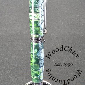 Może przedstawiać: Ręcznie wykonany długopis z żywym zielono-białym marmurkowym wzorem, zaakcentowany srebrnymi metalowymi elementami. Długopis jest prezentowany na srebrnym stojaku na długopisy. Widoczny jest tekst "WoodChux Est. 1999".