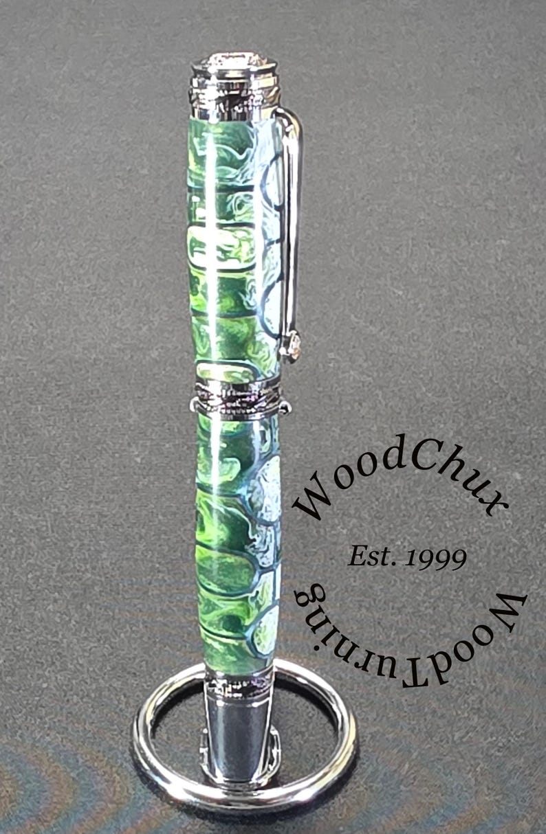 Może przedstawiać: Ręcznie wykonany długopis z żywym zielono-białym marmurkowym wzorem, zaakcentowany srebrnymi metalowymi elementami. Długopis jest prezentowany na srebrnym stojaku na długopisy. Widoczny jest tekst "WoodChux Est. 1999".